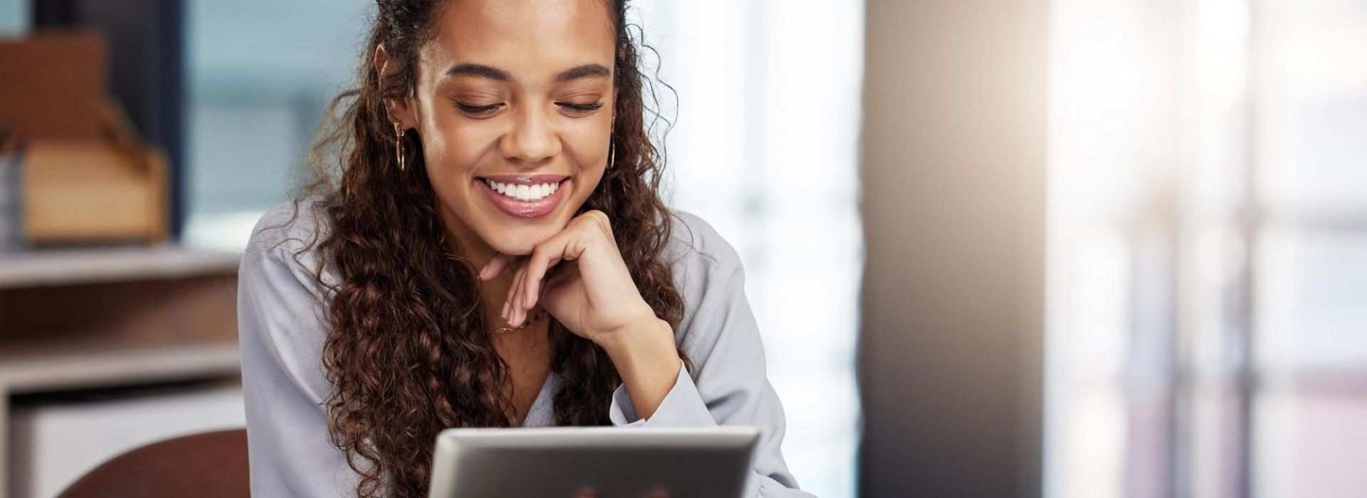 Glückliche Frau am Bürotisch blickt auf Tablet, welches in der Hand gehalten wird.